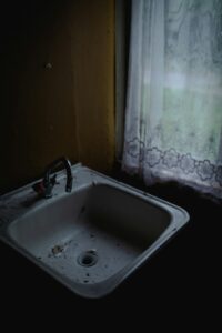 A homeowner checking under a kitchen sink for leaks, with cleaning supplies and visible plumbing pipes.