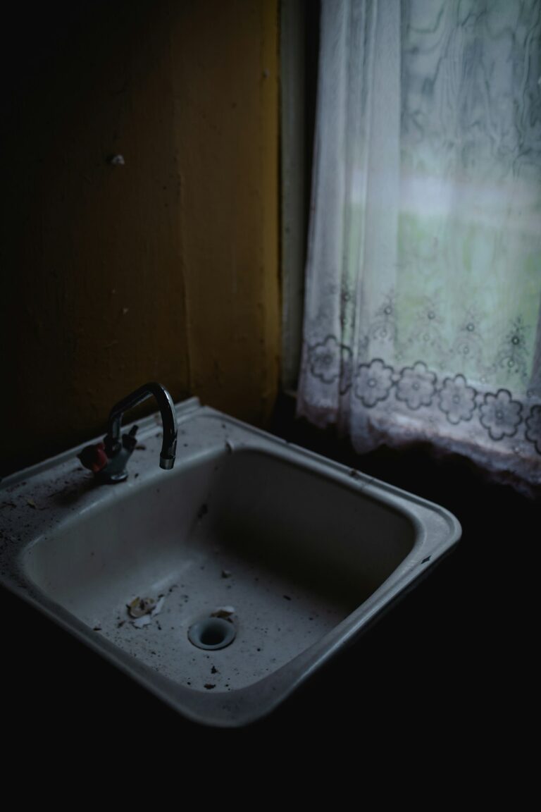A homeowner checking under a kitchen sink for leaks, with cleaning supplies and visible plumbing pipes.