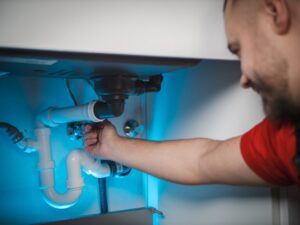 A homeowner kneeling under a kitchen sink inspecting pipes with a flashlight.