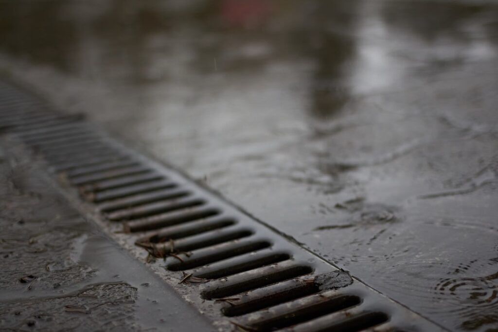 A gloved hand holding a plumbing snake near a residential floor drain with standing water.