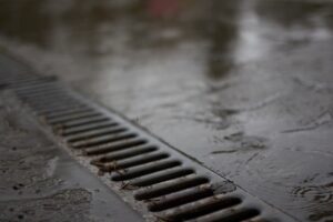 A gloved hand holding a plumbing snake near a residential floor drain with standing water.
