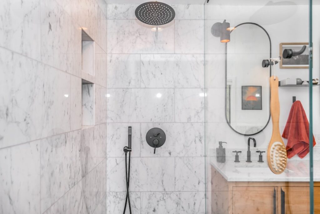 A person testing hot water flow in a household shower with tiled walls and a standard showerhead.