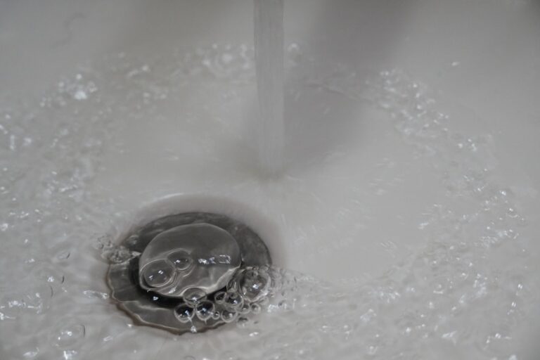 A person cleaning a bathroom sink drain with a brush and gloves, with water running into the basin.