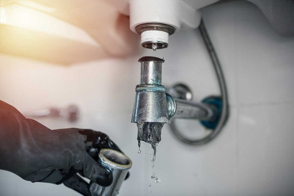 A homeowner examines a corroded water pipe beneath a kitchen sink with a flashlight.