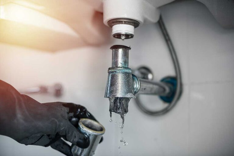 A homeowner examines a corroded water pipe beneath a kitchen sink with a flashlight.